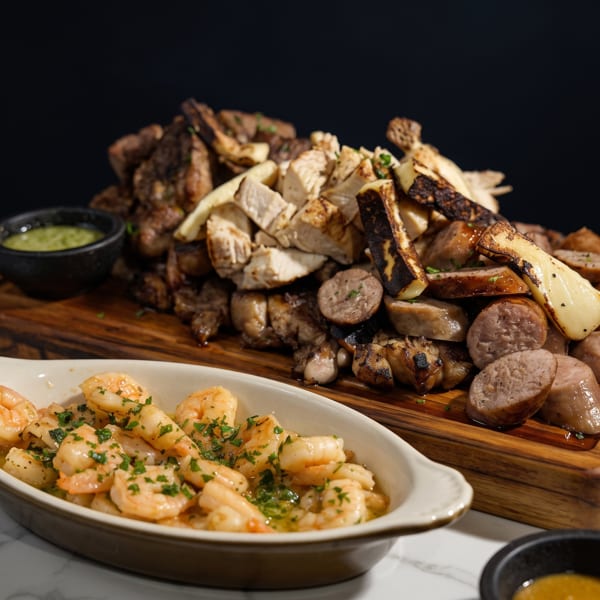 Grilled meats, sausages, and mushrooms on a wooden board, with a side of garlic shrimp.