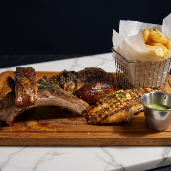 Grilled ribs, chicken, sausage, and fries with green sauce on a wooden board.